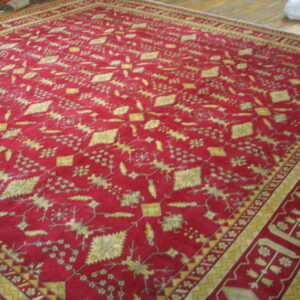 Deep red rug with an overall repeating gold geometric and botanical pattern on a wooden floor.