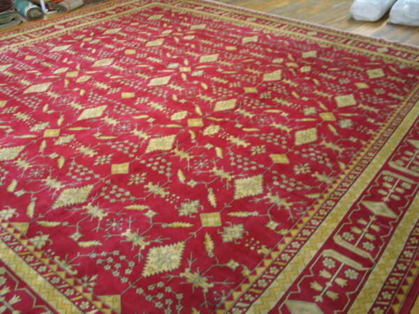 Deep red rug with an overall repeating gold geometric and botanical pattern on a wooden floor.