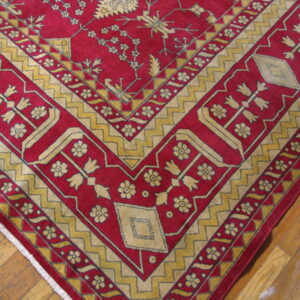Red rug featuring stylized gold floral designs and zigzag borders resting on wood plank flooring.