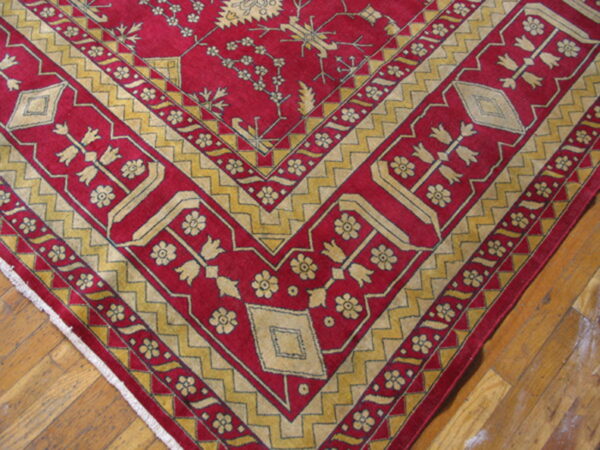 Red rug featuring stylized gold floral designs and zigzag borders resting on wood plank flooring.