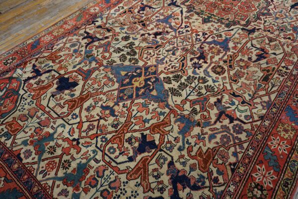 Angled view of a patterned rug with cream ground, red, and blue motifs on worn wood planks.