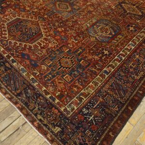 Red and navy high-detail geometric rug covers a corner of distressed light brown wood flooring.