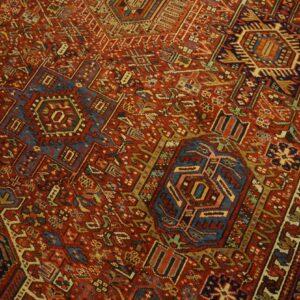 Complex geometric rug pattern featuring rich brick red, navy blue, cream, and gold details.