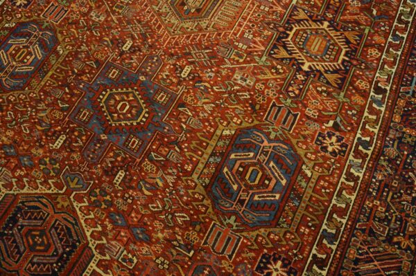 Complex geometric rug pattern featuring rich brick red, navy blue, cream, and gold details.