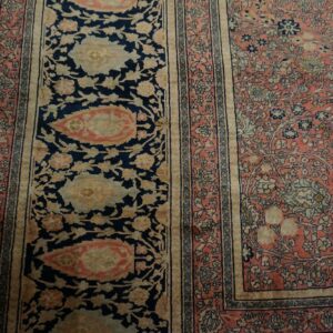 Close-up of a floral rug with navy blue border panels and a salmon pink field on wooden flooring.