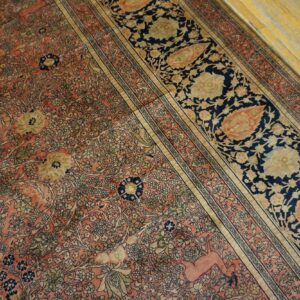 Intricate low-pile rug in coral, navy, and gold featuring floral motifs and a deer figure on worn wood flooring.