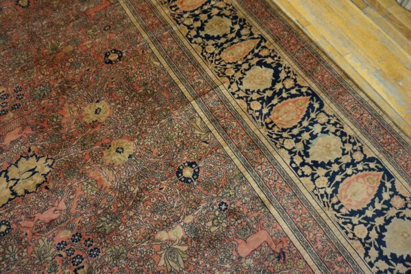 Intricate low-pile rug in coral, navy, and gold featuring floral motifs and a deer figure on worn wood flooring.