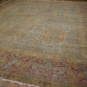 Large antique rug in faded teal, gold, and maroon floral pattern resting on wood flooring.