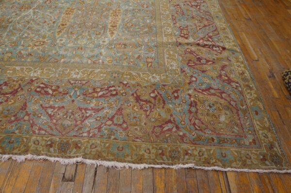Low-pile rug in faded teal, gold, and red traditional patterns with white fringe on wood floor.