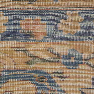 Close-up view of a low-pile rug showing a traditional pattern in tan, dark blue, rust, and brown accents.