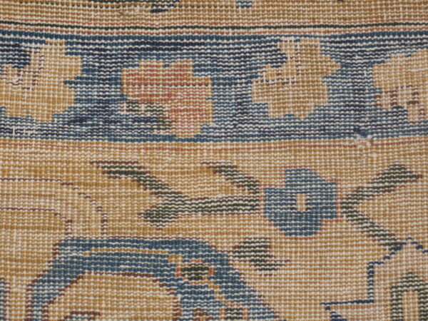 Close-up view of a low-pile rug showing a traditional pattern in tan, dark blue, rust, and brown accents.