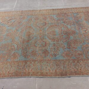 Large ornate low-pile rug featuring a distressed rust and light blue floral design over concrete flooring.