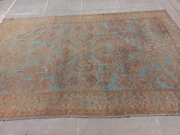 Large ornate low-pile rug featuring a distressed rust and light blue floral design over concrete flooring.