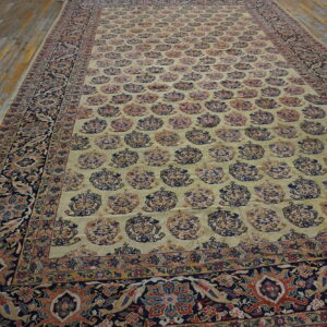 Traditional rug with a beige-yellow field, repeating boteh motifs, and a navy floral border on wood floors.