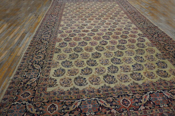 Traditional rug with a beige-yellow field, repeating boteh motifs, and a navy floral border on wood floors.