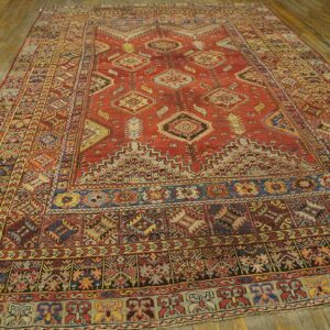 Low pile rug in rust red with dense blue and yellow geometric patterns on wooden floorboards.