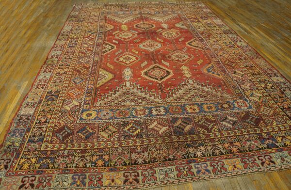 Low pile rug in rust red with dense blue and yellow geometric patterns on wooden floorboards.