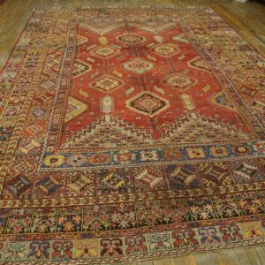 Faded red rug with complex geometric medallions and multi-color borders covers worn light-toned wood flooring.