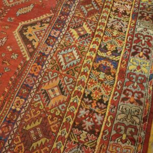 Close-up of a rust-red rug with intricate multi-color tribal borders on textured wood plank flooring.