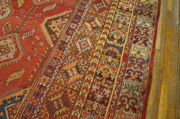 Close-up of a rust-red rug with intricate multi-color tribal borders on textured wood plank flooring.
