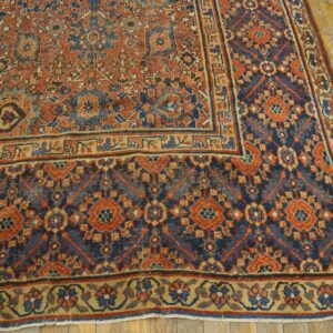 Low-pile rug in rust, blue, and tan with geometric patterns displayed on worn hardwood flooring.