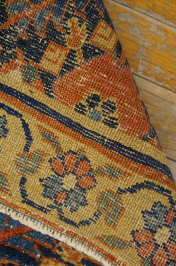 Detail of a low-pile rug showing navy, orange, and gold ornate motifs against yellow distressed wood.