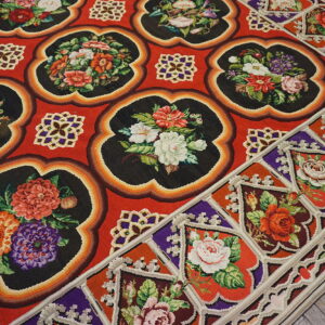 Red rug with black scalloped panels displaying multicolored floral designs on gray wooden flooring.