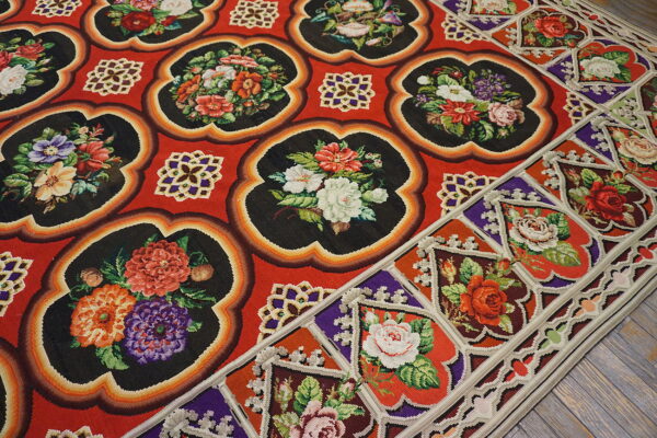 Red rug with black scalloped panels displaying multicolored floral designs on gray wooden flooring.