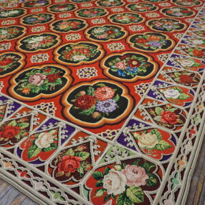Intricate red tapestry rug featuring repeating black floral medallions and an elaborate rose border on wood planks.