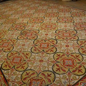Low-pile rug displaying an intricate quatrefoil pattern in cream, red, olive green, and gold on wood flooring.