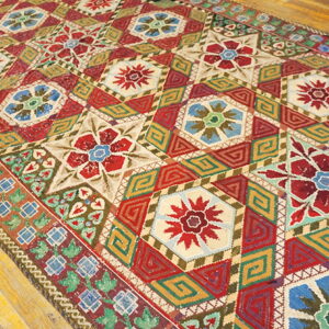 Intricately patterned rug in red, gold, blue, and cream sits angled on wood plank flooring.