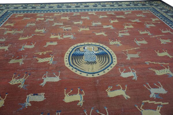 Textured red field rug featuring scattered pale animal figures, a central blue bird medallion, and wide blue borders.