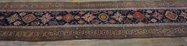Narrow runner rug with a navy blue field, repeated geometric medallions, and detailed floral borders on wood flooring.