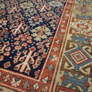 A detailed view of a navy, red, and beige low-pile rug showing complex geometric motifs on wood floor.
