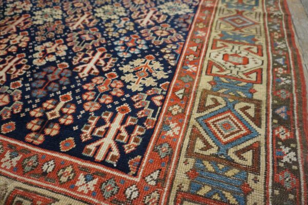 A detailed view of a navy, red, and beige low-pile rug showing complex geometric motifs on wood floor.