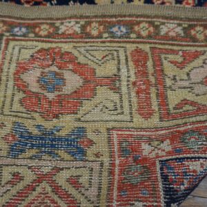 A detailed view of a traditional rug border in red, gold, and blue resting on weathered wood.