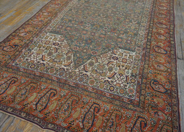 Floral patterned rug with teal and rust colors, featuring a border of large paisley motifs on wood flooring.