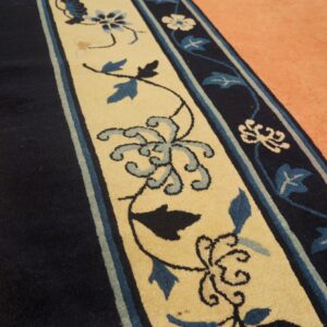 A navy and beige border rug with blue florals rests against wood and peach carpet.
