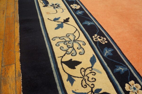 A navy and beige border rug with blue florals rests against wood and peach carpet.