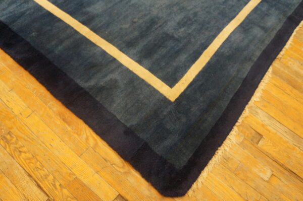 Corner of a medium-pile blue rug featuring navy and cream concentric borders on bright hardwood flooring.