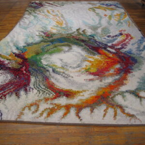 Textured white area rug featuring a dense, multicolored abstract vortex design, displayed on plank wood flooring.