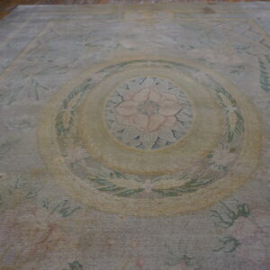 A distressed low-pile rug featuring a pale gray-green, blush pink, and yellow floral medallion on a wood floor.