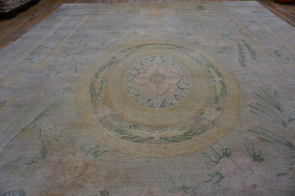 A distressed low-pile rug featuring a pale gray-green, blush pink, and yellow floral medallion on a wood floor.