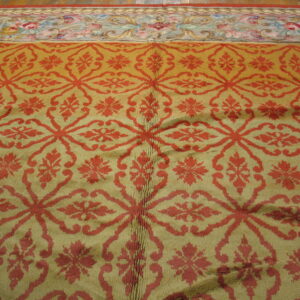 Muted gold rug featuring a repeating red ogee lattice pattern and a multicolored floral border on a wood floor.