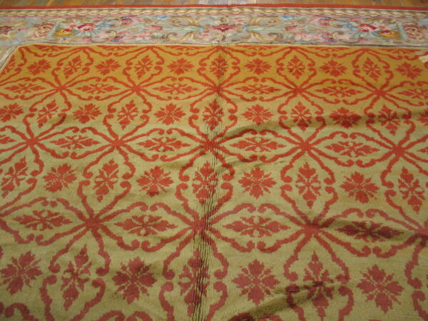Muted gold rug featuring a repeating red ogee lattice pattern and a multicolored floral border on a wood floor.
