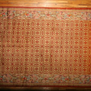 Golden-tan rug with red trellis pattern and detailed multi-color floral border on a hardwood floor.