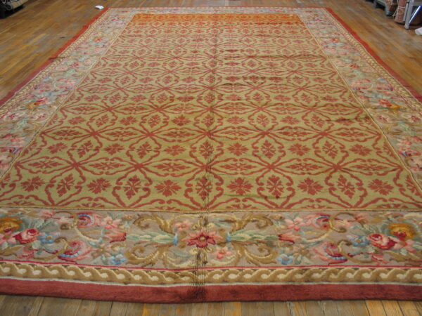 Large rug with a red geometric pattern on an olive field and wide multicolored floral borders on wood floor.