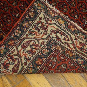Close-up of a low-pile red rug folded over, revealing the multicolored geometric borders on wood planks.