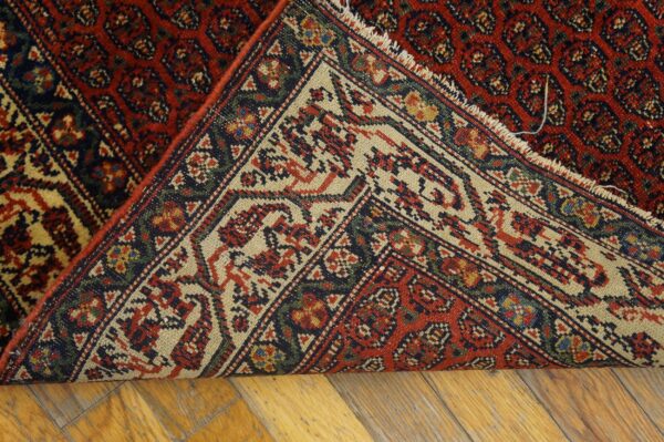 Close-up of a low-pile red rug folded over, revealing the multicolored geometric borders on wood planks.