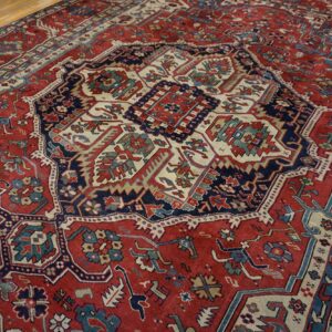 Densely patterned red rug with complex geometric motifs in navy, cream, and teal on wood flooring.